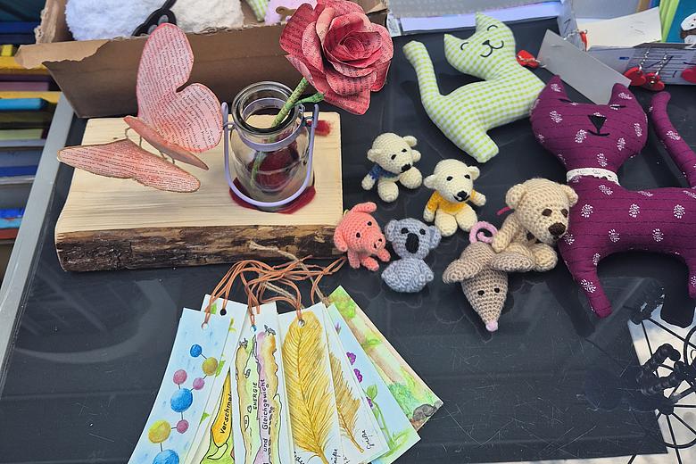 Das Foto zeigt einen Tisch auf dem Handarbeiten liegen. Es sind zu sehen bemalte Lesezeichen, gestrickte und genähte Tiere und ein Schmetterling und eine Rose aus Origami.