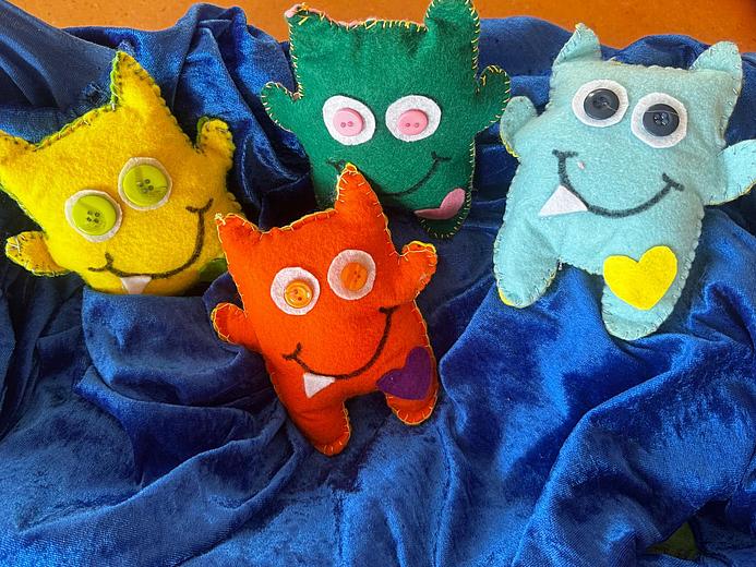 Vier bunte Stofftiere in Monsterform (gelb, orange, grün und hellblau). Sie liegen auf einem dunkelblauen Untergrund.