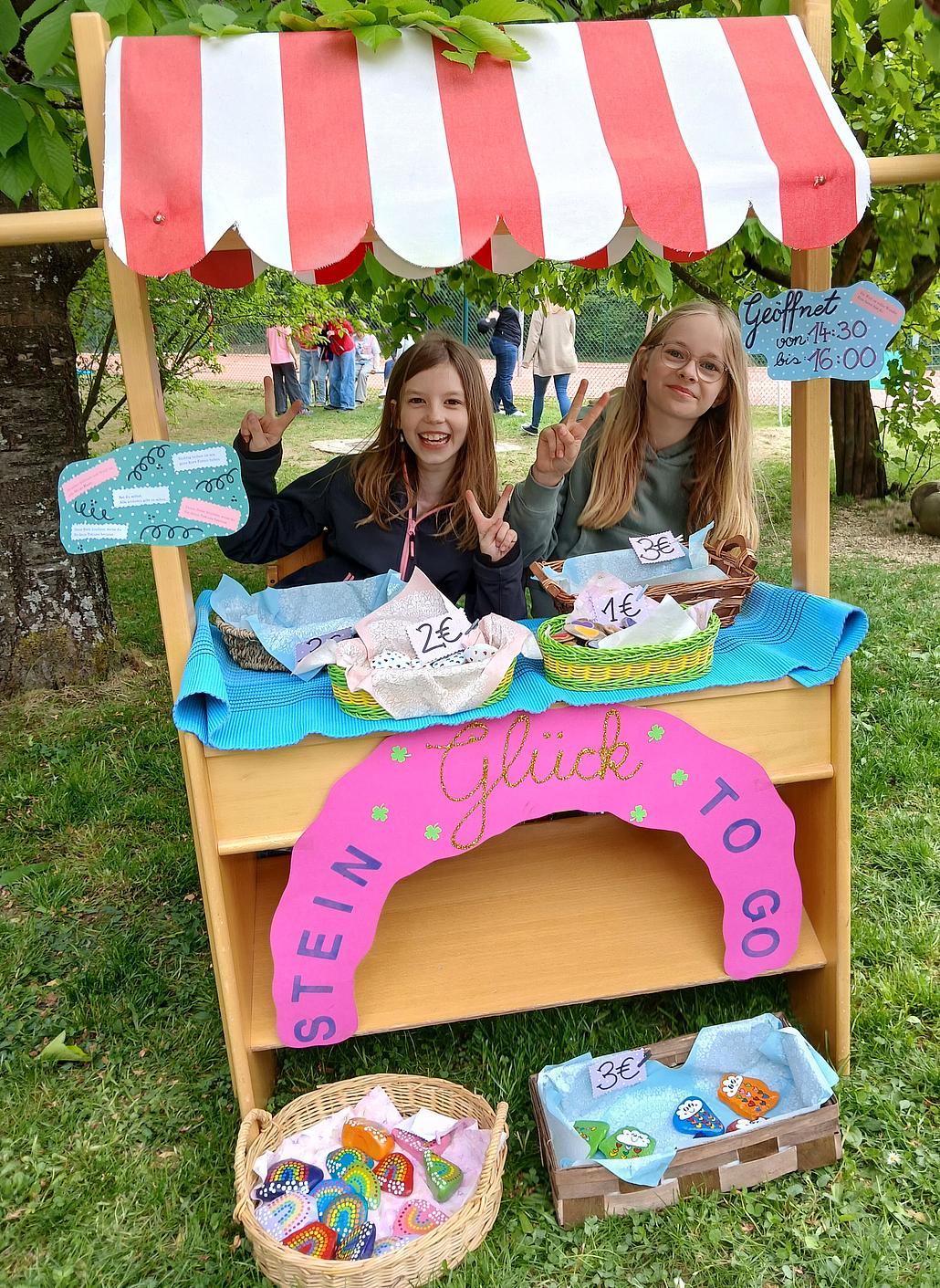 Zwei Mädchen sitzen lachend hinter einem kleinen, hölzernen Verkaufsstand im Freien. Der Stand hat ein rot-weiß gestreiftes Stoffdach und ist mit bunten Schildern dekoriert. Auf einem pinken Schild unter der Theke steht in großen Buchstaben „Glückstein to go“. Auf dem Tisch sind drei Körbe mit bunt bemalten Glückssteinen, die jeweils mit einem Preisschild (1 €, 2 € und 3 €) versehen sind. Ein zusätzlicher Korb mit weiteren Steinen steht auf dem Rasen vor dem Stand. Ein handgeschriebenes Schild oben rechts am Stand informiert über die Öffnungszeiten: „Geöffnet von 14:30 bis 16:00“. Hinter dem Stand stehen Bäume, und im Hintergrund sind weitere Menschen zu sehen, vermutlich auf einem Fest oder Markt. Ein Erwachsener beugt sich rechts ins Bild. Die Atmosphäre ist fröhlich und lebendig.
