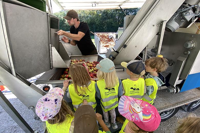 Die Kinder schauen einen Mann zu, welcher gerade Äpfel in die Apfelpresse verlädt.