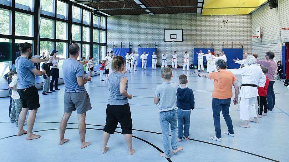 Menschen stehen in einem großen Kreis in einer Sporthalle. Sie machen gemeinsam Karateübungen. Hinten zu sehen sind Menschen in weißen Karateanzügen. Vorne, mit dem Rücken zur Kamera, stehen Erwachsene und Kinder in Alltagskleidung.