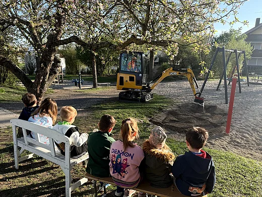 Sieben Kinder sind von hinten zu sehen. Sie sitzen auf zwei Bänken, die unter einem blühenden Apfelbaum stehen. Sie schauen dabei zu, wie ein Minibagger Kies schaufelt. Im Hintergrund sind eine Schaukel und weitere Spielgeräte eines Spielplatzes zu sehen.