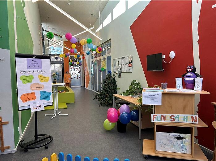 Zu sehen ist ein heller, freundlicher Flur der Kindertageseinrichtung. Der Raum ist farbenfroh gestaltet: Die Wände sind in Grün, Orange und Rot gestrichen. Von der Decke hängen bunte Papierkugeln und Dekorationen. Auf dem Boden stehen mehrere bunte Luftballons.  Links im Bild befindet sich ein Aufsteller mit einem Plakat zum „Tag der offenen Tür“. Darauf ist ein Programm für Kinder angekündigt. Daneben stehen kleine Sitzmöbel in Grün. Im hinteren Bereich des Flurs sind Glastüren und Fenster zu sehen, durch die Tageslicht einfällt.  Rechts im Vordergrund steht ein Empfangstisch aus Holz. Darauf liegen Informationsmaterialien und eine kleine Pflanze. An der Front des Tisches ist ein Schild mit der Aufschrift „Fundsachen“ angebracht. In einem offenen Fach darunter liegen Zeichnungen von Kindern. An der Wand dahinter hängt ein Bildschirm sowie ein schwarzes Stoffmaskottchen.