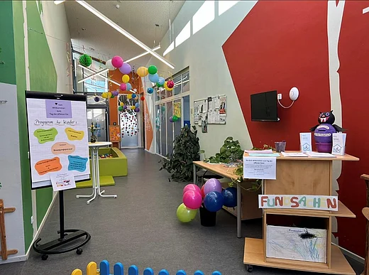Zu sehen ist ein heller, freundlicher Flur der Kindertageseinrichtung. Der Raum ist farbenfroh gestaltet: Die Wände sind in Grün, Orange und Rot gestrichen. Von der Decke hängen bunte Papierkugeln und Dekorationen. Auf dem Boden stehen mehrere bunte Luftballons.  Links im Bild befindet sich ein Aufsteller mit einem Plakat zum „Tag der offenen Tür“. Darauf ist ein Programm für Kinder angekündigt. Daneben stehen kleine Sitzmöbel in Grün. Im hinteren Bereich des Flurs sind Glastüren und Fenster zu sehen, durch die Tageslicht einfällt.  Rechts im Vordergrund steht ein Empfangstisch aus Holz. Darauf liegen Informationsmaterialien und eine kleine Pflanze. An der Front des Tisches ist ein Schild mit der Aufschrift „Fundsachen“ angebracht. In einem offenen Fach darunter liegen Zeichnungen von Kindern. An der Wand dahinter hängt ein Bildschirm sowie ein schwarzes Stoffmaskottchen.