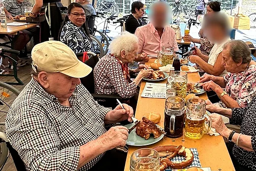 Mehrere ältere Menschen sitzen an festlich gedeckten Biertischen in einem Festzelt und essen gemeinsam bayerische Spezialitäten. Die Stimmung wirkt gesellig und entspannt.