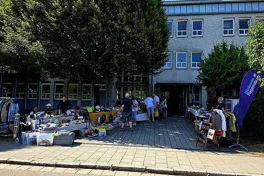 Eine Außenansicht des Johann-Hinrich-Wichern-Haus. Vor dem Haus stehen große Bäume und es sind Flohmarktstände aufgebaut. Einige Menschen sind zu sehen, ein Teil verkauft Waren, andere sind zum Stöbern gekommen.