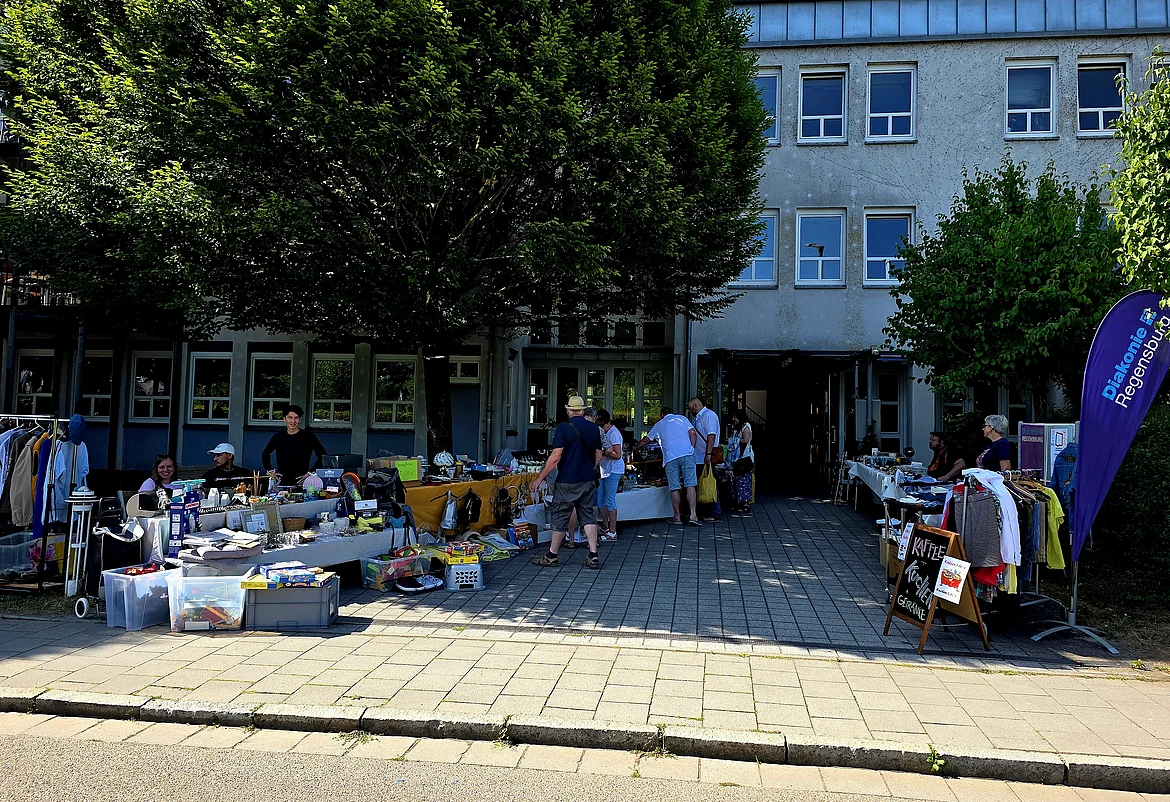 Eine Außenansicht des Johann-Hinrich-Wichern-Haus. Vor dem Haus stehen große Bäume und es sind Flohmarktstände aufgebaut. Einige Menschen sind zu sehen, ein Teil verkauft Waren, andere sind zum Stöbern gekommen.