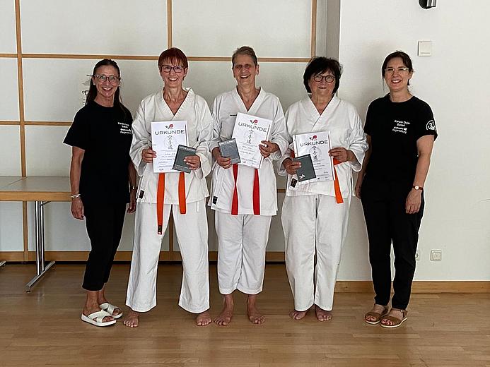 Das Bild zeigt drei Frauen in Karatekleidung, die ihre Uhrkunden zur erfolgreich absolvierten Karateprüfung in den Händen halten. Links und rechts stehen die beiden Trainierinnen in schwarzer Sportkleidung.