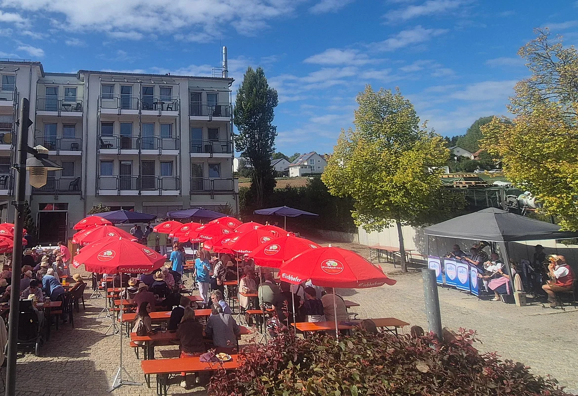 Das Bild zeigt einen Platz auf dem Biertische und Bierbänke unter roten Sonnenschirmen stehen. Im Hintergrund links ist ein mehrstöckiges, helles Gebäude zu sehen. Auf der rechten Seite sieht man Bäume und einen dunklen Pavillon. Darunter sitzt eine Blaskapelle. 