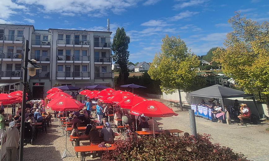 Das Bild zeigt einen Platz auf dem Biertische und Bierbänke unter roten Sonnenschirmen stehen. Im Hintergrund links ist ein mehrstöckiges, helles Gebäude zu sehen. Auf der rechten Seite sieht man Bäume und einen dunklen Pavillon. Darunter sitzt eine Blaskapelle. 