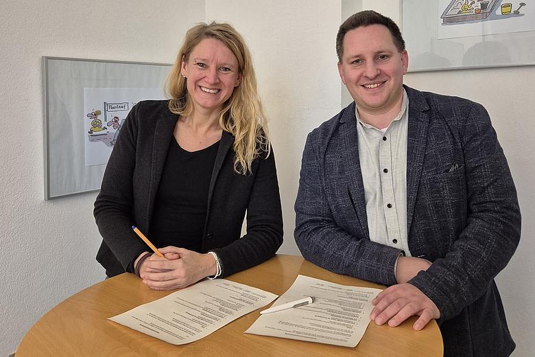 Zwei erwachsene Personen stehen hinter einem runden Holztisch und lächeln in die Kamera. Auf dem Tisch liegen zwei unterschriebene Dokumente sowie ein Stift. Die Person links trägt dunkle Kleidung und hält einen Stift in der Hand, die Person rechts trägt ein helles Hemd mit dunklem Sakko. Beide scheinen gerade eine Vertragsunterzeichnung abgeschlossen zu haben. Im Hintergrund ist eine helle Wand mit gerahmten Illustrationen zu sehen.