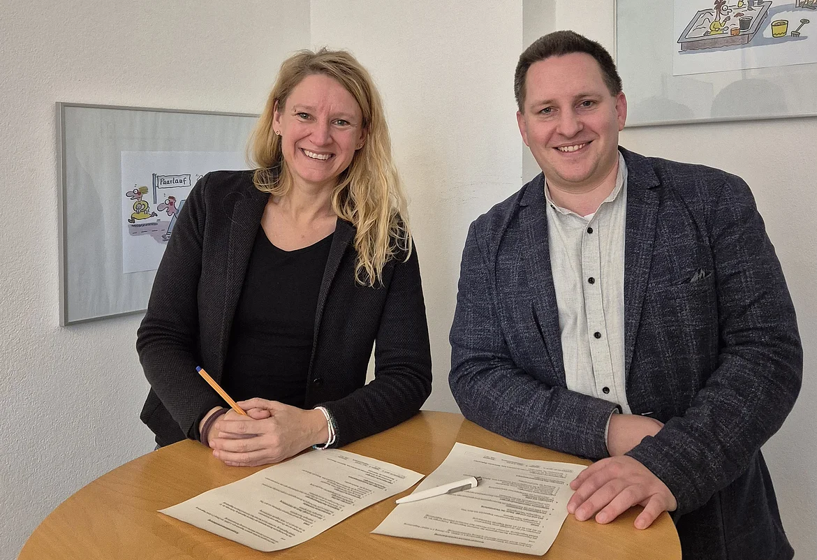 Zwei erwachsene Personen stehen hinter einem runden Holztisch und lächeln in die Kamera. Auf dem Tisch liegen zwei unterschriebene Dokumente sowie ein Stift. Die Person links trägt dunkle Kleidung und hält einen Stift in der Hand, die Person rechts trägt ein helles Hemd mit dunklem Sakko. Beide scheinen gerade eine Vertragsunterzeichnung abgeschlossen zu haben. Im Hintergrund ist eine helle Wand mit gerahmten Illustrationen zu sehen.