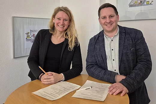 Zwei erwachsene Personen stehen hinter einem runden Holztisch und lächeln in die Kamera. Auf dem Tisch liegen zwei unterschriebene Dokumente sowie ein Stift. Die Person links trägt dunkle Kleidung und hält einen Stift in der Hand, die Person rechts trägt ein helles Hemd mit dunklem Sakko. Beide scheinen gerade eine Vertragsunterzeichnung abgeschlossen zu haben. Im Hintergrund ist eine helle Wand mit gerahmten Illustrationen zu sehen.