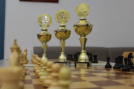 Zu sehen sind drei goldene Pokale in unterschiedlichen Größen, aufgereiht von klein nach groß. Außenherum sind etwas verschwommen Schachfiguren aus Holz erkennbar, die auf einem Schachbrett stehen.