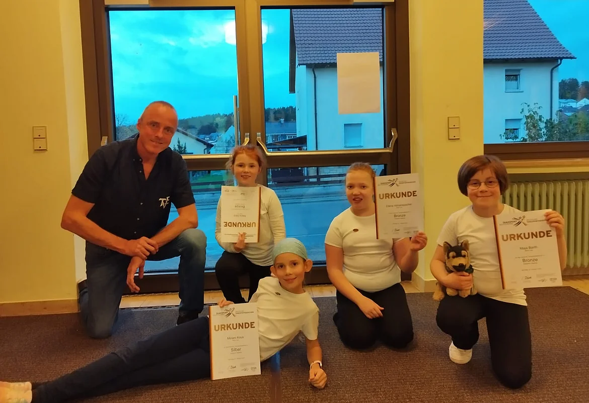 Vier Kinder und ein Erwachsener posieren in einem hellen Raum vor großen Fenstern. Die Kinder tragen weiße T-Shirts und dunkle Hosen. Drei von ihnen halten Urkunden in der Hand, eines hält zusätzlich ein Stofftier. Ein Mädchen liegt vorn am Boden und zeigt ebenfalls stolz ihre Urkunde. Der Erwachsene kniet links neben der Gruppe und lächelt. Draußen ist es bereits dämmrig, man sieht Häuser und einen blauen Abendhimmel.