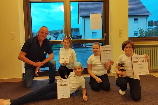 Vier Kinder und ein Erwachsener posieren in einem hellen Raum vor großen Fenstern. Die Kinder tragen weiße T-Shirts und dunkle Hosen. Drei von ihnen halten Urkunden in der Hand, eines hält zusätzlich ein Stofftier. Ein Mädchen liegt vorn am Boden und zeigt ebenfalls stolz ihre Urkunde. Der Erwachsene kniet links neben der Gruppe und lächelt. Draußen ist es bereits dämmrig, man sieht Häuser und einen blauen Abendhimmel.