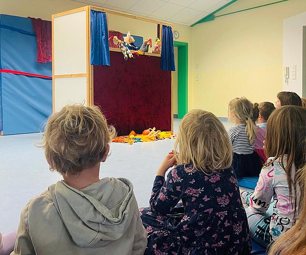 Kinderhaus Thalmassing_Kasperltheater 2025 Mehrere Kinder sitzen im Kindergarten auf dem Boden in einem hellen Raum und schauen gespannt auf ein Kasperltheater. Vor ihnen steht eine kleine Bühne mit rotem Vorhang und blauen Seitenteilen. Hinter der Bühne sind Handpuppen zu sehen, die gerade eine Szene spielen. Auf dem Boden vor der Bühne liegen bunte Stofftiere. Die Stimmung wirkt fröhlich und konzentriert.