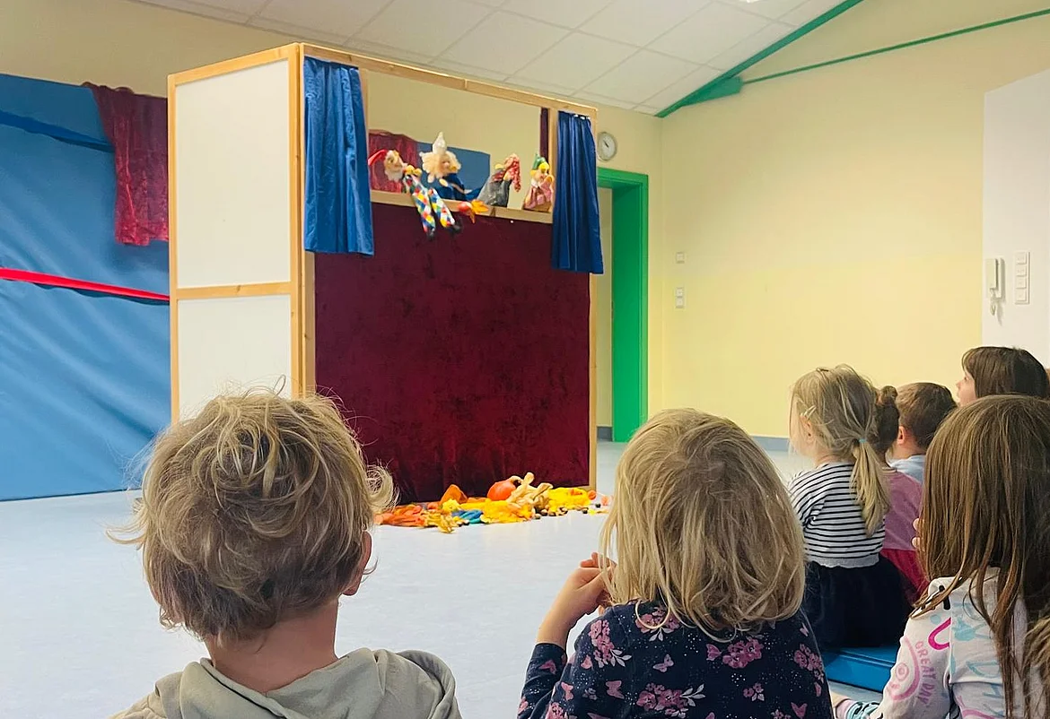 Mehrere Kinder sitzen im Kindergarten auf dem Boden in einem hellen Raum und schauen gespannt auf ein Kasperltheater. Vor ihnen steht eine kleine Bühne mit rotem Vorhang und blauen Seitenteilen. Hinter der Bühne sind Handpuppen zu sehen, die gerade eine Szene spielen. Auf dem Boden vor der Bühne liegen bunte Stofftiere. Die Stimmung wirkt fröhlich und konzentriert.