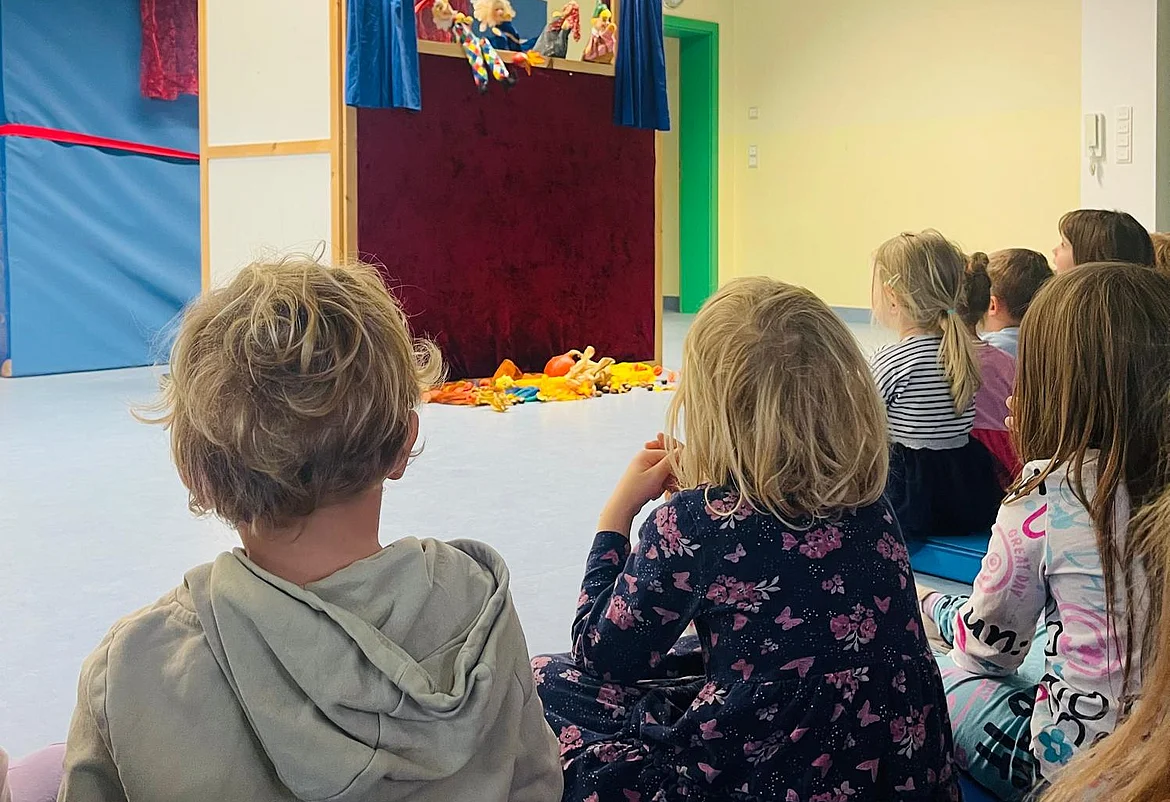 Mehrere Kinder sitzen im Kindergarten auf dem Boden in einem hellen Raum und schauen gespannt auf ein Kasperltheater. Vor ihnen steht eine kleine Bühne mit rotem Vorhang und blauen Seitenteilen. Hinter der Bühne sind Handpuppen zu sehen, die gerade eine Szene spielen. Auf dem Boden vor der Bühne liegen bunte Stofftiere. Die Stimmung wirkt fröhlich und konzentriert.