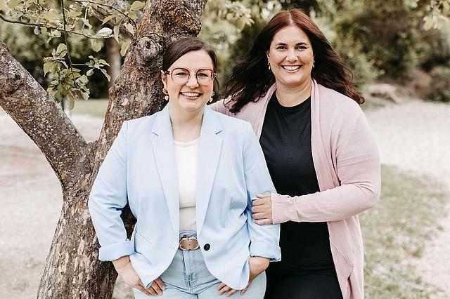 Das Foto zeigt zwei Frauen. Julia Berghammer, die Leitung des Kinderhauses St. Nikolaus, und Talia Kastler, die stellvertretende Leitung. Sie stehen im Grünen, unter einen Baum und lächeln freundlich.