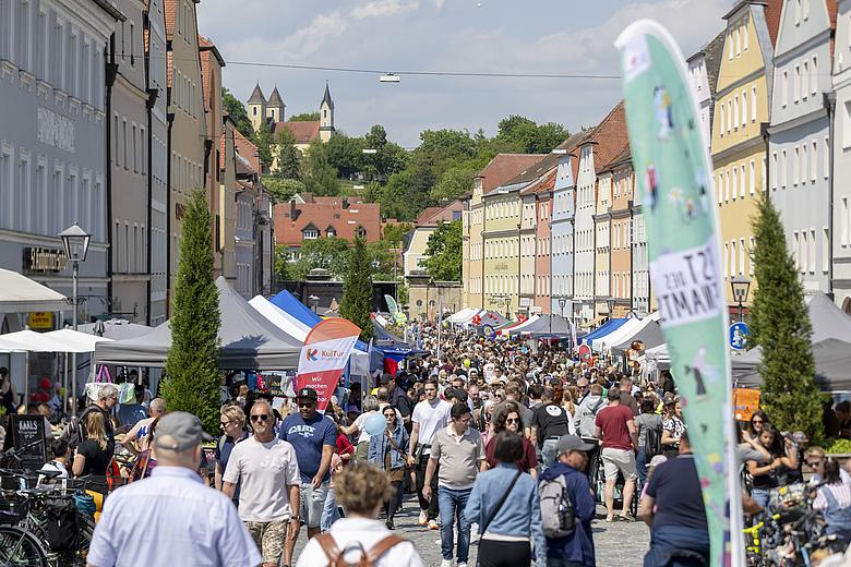 Belebte Straßenszene bei einer öffentlichen Veranstaltung in der Altstadt von Regensburg. Zahlreiche Menschen bewegen sich zwischen Informationsständen und Fahnen entlang einer breiten Straße mit historischen Gebäuden. Im Hintergrund sind Grünflächen und Bauwerke auf einem Hügel zu sehen. Sonniges Wetter und eine offene, lebendige Atmosphäre prägen das Bild.