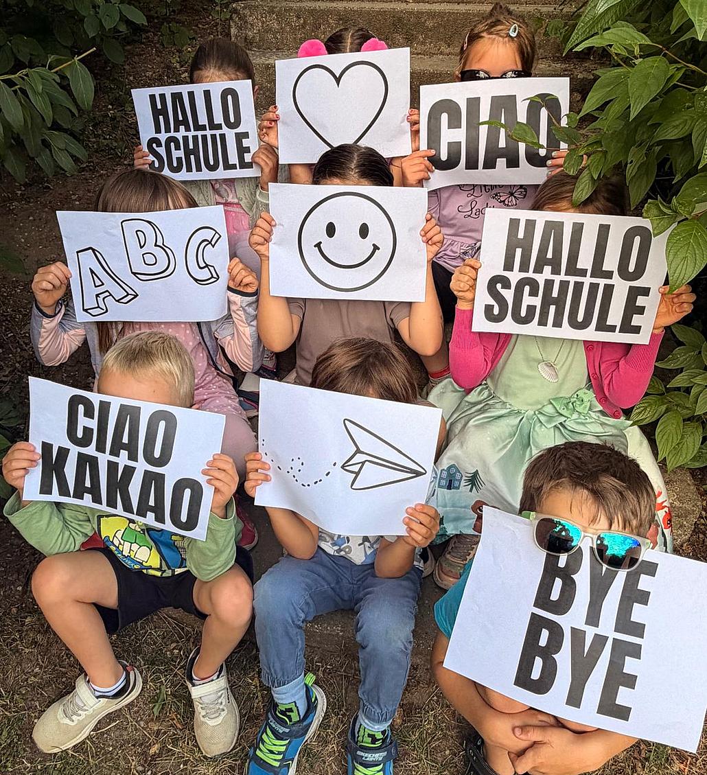 Neun Kinder sitzen auf einer Treppe, jeweils drei in einer Reihe. Ihre Gesichter sind hinter Papierbögen versteckt auf denen jeweils ein anderes Motiv oder Spruch zu sehen ist. Darauf sind: ein Herz, ein Smiley, ein Pfeil, Hallo Schule, Ciao, Ciao Kakao und Bye Bye.
