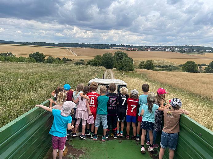 Ungefähr 15 Kinder sind von hinten zu sehen. Sie stehen auf dem Anhänger eines Traktors und fahren über die Felder. Im Hintergrund ist die Weite zu sehen mit vielen Feldern, größtenteils Kornfelder. 
