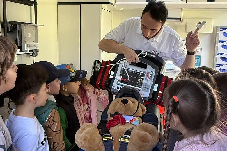 Ein Mitarbeiter des Rettungsdienstes erklärt einer Gruppe von Kindern die Funktion eines medizinischen Geräts in einem Rettungswagen. Vor ihm liegt ein Stofftier angeschnallt auf einer Trage, während die Kinder aufmerksam zuhören. Die Szene vermittelt spielerisch Wissen über Erste Hilfe, Notfallversorgung und die Arbeit im Rettungsdienst.