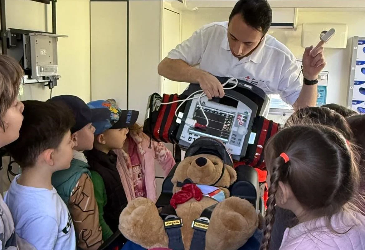 Ein Mitarbeiter des Rettungsdienstes erklärt einer Gruppe von Kindern die Funktion eines medizinischen Geräts in einem Rettungswagen. Vor ihm liegt ein Stofftier angeschnallt auf einer Trage, während die Kinder aufmerksam zuhören. Die Szene vermittelt spielerisch Wissen über Erste Hilfe, Notfallversorgung und die Arbeit im Rettungsdienst.