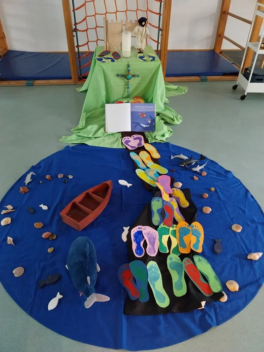 Auf dem Foto ist eine kindgerechte und kreativ gestaltete Erzähl- und Gebetslandschaft im Kindergarten zu sehen. Sie stellt die biblische Geschichte von Jona dar. In der Mitte liegt ein großes rundes blaues Tuch auf dem Boden – es symbolisiert das Meer. Darauf sind Muscheln, Steine, kleine Holzfische und ein großer blauer Stoffwal verteilt, der an den Fisch erinnert, der Jona verschluckt.  Ein kleines rotes Holzboot schwimmt im „Meer“. Von dort führt ein schwarzer Stoffweg mit vielen bunten Fußabdrücken aus Papier in Richtung eines Altars. Der Altar ist mit grünem Stoff bedeckt und zeigt eine Jesusfigur, eine große Kerze, ein Kreuz aus bunten Perlen und weitere bunte Fußspuren. Davor liegt ein geöffnetes Bilderbuch mit einer Szene aus der Jona-Geschichte: Jona fällt ins Wasser, und der Fisch taucht auf.  Diese Gestaltung lädt die Kinder ein, die Geschichte von Jona spielerisch zu entdecken und über Mut, Angst und Vertrauen nachzudenken.
