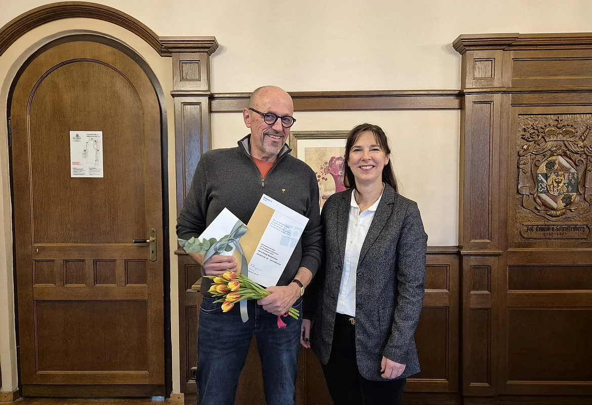 Zwei Personen stehen nebeneinander in einem holzgetäfelten Raum. Die Person links hält einen Blumenstrauß und eine Urkunde, beide lächeln in die Kamera.