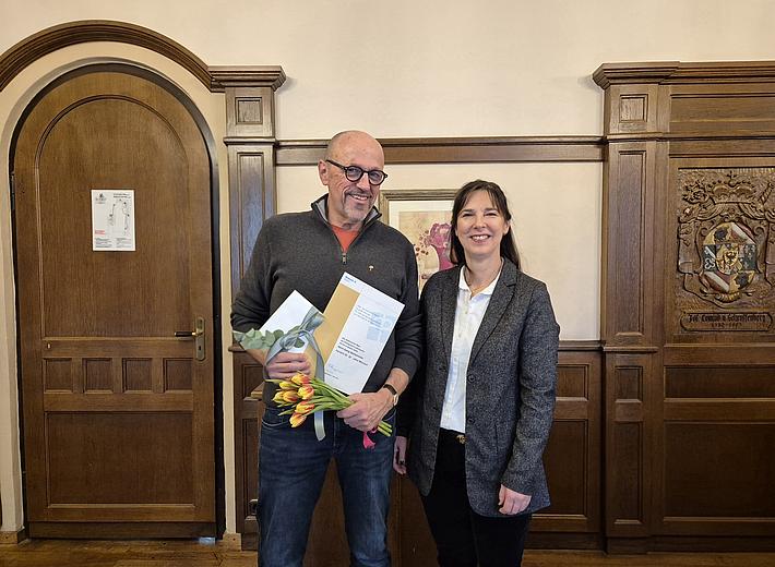 Zwei Personen stehen nebeneinander in einem holzgetäfelten Raum. Die Person links hält einen Blumenstrauß und eine Urkunde, beide lächeln in die Kamera.