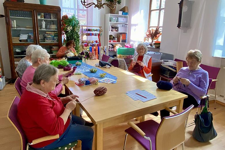 Um einen Tisch sitzen sechs Seniorinnen. Drei von ihnen stricken, zwei häkeln und eine drapiert Kunstblumen auf einen Strohhut. Die Atmosphäre wirkt gesellig, durch die Fenster scheint die Sonne.
