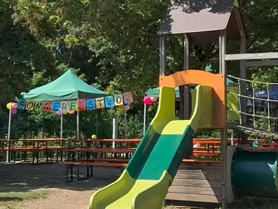 Im Garten des Kindergartens sind Biertische unter Sonnenschirmen aufgestellt. Eine bunte Girlande zeigt den Schriftzug "Sommerfest 2026". Im Vordergrund ist eine Rusche zu sehen.