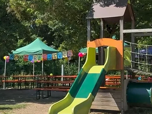 Im Garten des Kindergartens sind Biertische unter Sonnenschirmen aufgestellt. Eine bunte Girlande zeigt den Schriftzug "Sommerfest 2026". Im Vordergrund ist eine Rusche zu sehen.