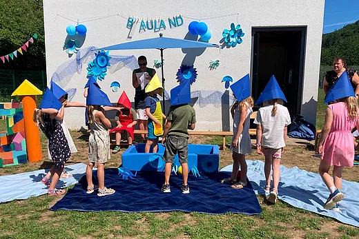 Kinder führen beim Sommerfest des Kinderhauses Tegernheim ein Theaterstück zum Thema „Blauland“ auf. Sie tragen bunte Papierhüte und stehen vor einer blau dekorierten Bühne im Außenbereich, während eine Betreuerin die Aufführung begleitet.