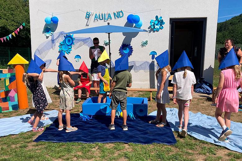 Kinder führen beim Sommerfest des Kinderhauses Tegernheim ein Theaterstück zum Thema „Blauland“ auf. Sie tragen bunte Papierhüte und stehen vor einer blau dekorierten Bühne im Außenbereich, während eine Betreuerin die Aufführung begleitet.