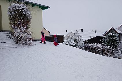 Zwei Kinder in bunter Winterkleidung spielen im Schnee auf einem kleinen Hügel im Garten des Kindergarten. Sie bewegen sich bergauf durch den tiefen Schnee. Im Hintergrund sind verschneite Dächer, Sträucher und Zäune eines Wohngebiets zu sehen. Die Umgebung wirkt ruhig und winterlich.