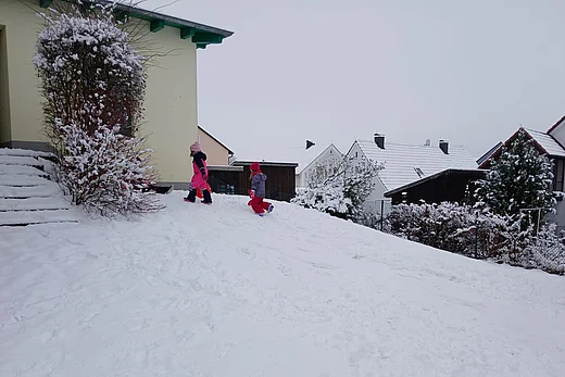 Zwei Kinder in bunter Winterkleidung spielen im Schnee auf einem kleinen Hügel im Garten des Kindergarten. Sie bewegen sich bergauf durch den tiefen Schnee. Im Hintergrund sind verschneite Dächer, Sträucher und Zäune eines Wohngebiets zu sehen. Die Umgebung wirkt ruhig und winterlich.