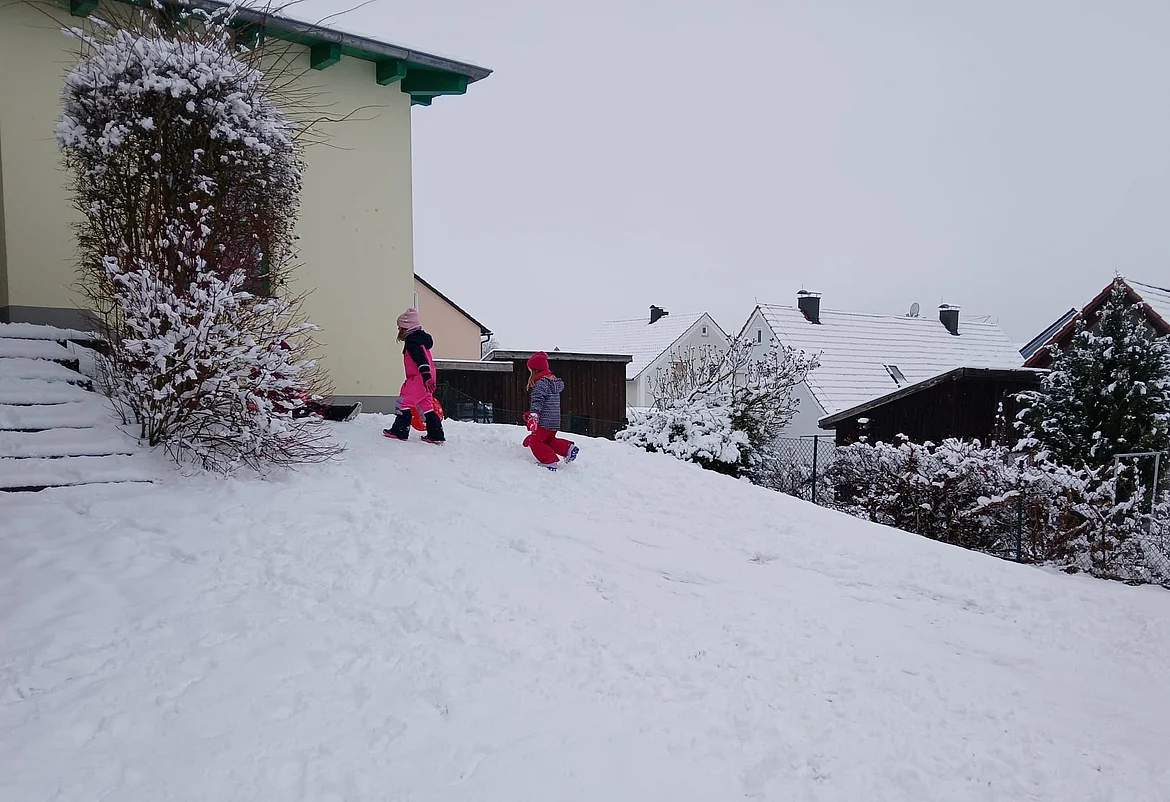 Zwei Kinder in bunter Winterkleidung spielen im Schnee im Garten des Kindergarten. Sie bewegen sich bergauf durch den tiefen Schnee. Im Hintergrund sind verschneite Dächer, Sträucher und Zäune eines Wohngebiets zu sehen. Die Umgebung wirkt ruhig und winterlich.