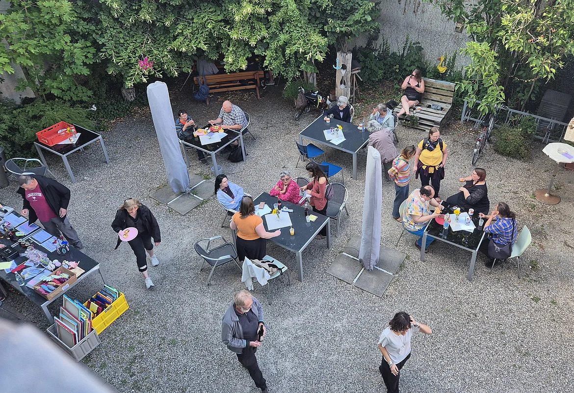 Der Garten des Café Insels von oben fotografiert. Es sitzen Menschen an Tischen, einige Stehen. Sie sind bunt gekleidet. Die Stimmung wirkt sehr positiv beim Sommerfest.
