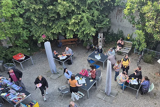 Der Garten des Café Insels von oben fotografiert. Es sitzen Menschen an Tischen, einige Stehen. Sie sind bunt gekleidet. Die Stimmung wirkt sehr positiv beim Sommerfest.