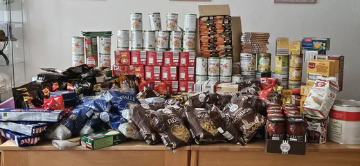 Auf einem großen Holztisch stehen zahlreiche haltbare Lebensmittel, ordentlich gestapelt. Zu sehen sind Konservendosen mit Eintöpfen sowie viele Packungen Pasta, Reis, Mehl, Brühe, Gewürze, Süßigkeiten und Saucen. 