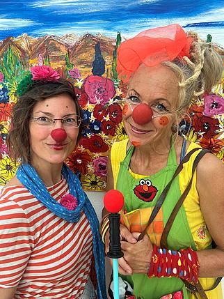 Zwei lächelnde Personen mit roten Clownsnasen und bunten Kostümen stehen vor einem farbenfrohen Gemälde mit Blumenwiese und Berglandschaft. Eine Person trägt ein rot-weiß gestreiftes Shirt und einen blauen Schal, die andere eine gelbe Kleidung mit grüner Schürze und auffälligem Haarschmuck.