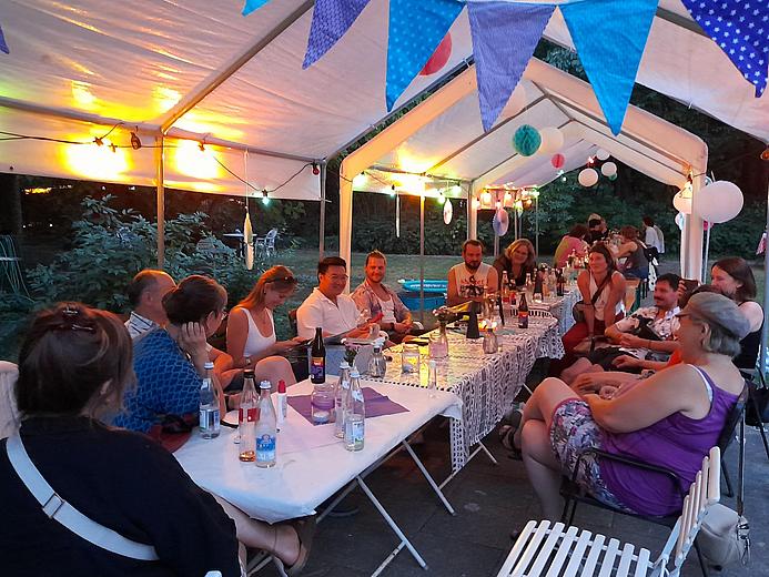 An einer langen Tafel sitzen Personen und unterhalten sich. Sie sitzen unter einem weißen Pavillon, der mit Lampen und Girlanden dekoriert ist. Es herrscht eine entspannte Stimmung in der Abenddämmerung.