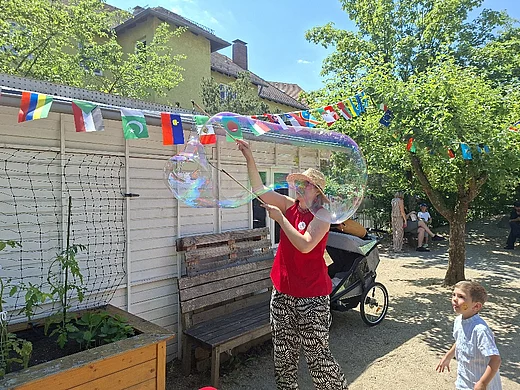 Eine Person erzeugt auf dem Sommerfest des Kindergartens mit Seifenblasenstäben eine große schillernde Seifenblase. Ein Kind beobachtet begeistert das Geschehen. Im Hintergrund sind bunte Länderflaggen, Bäume und weitere Besucherinnen und Besucher zu sehen.