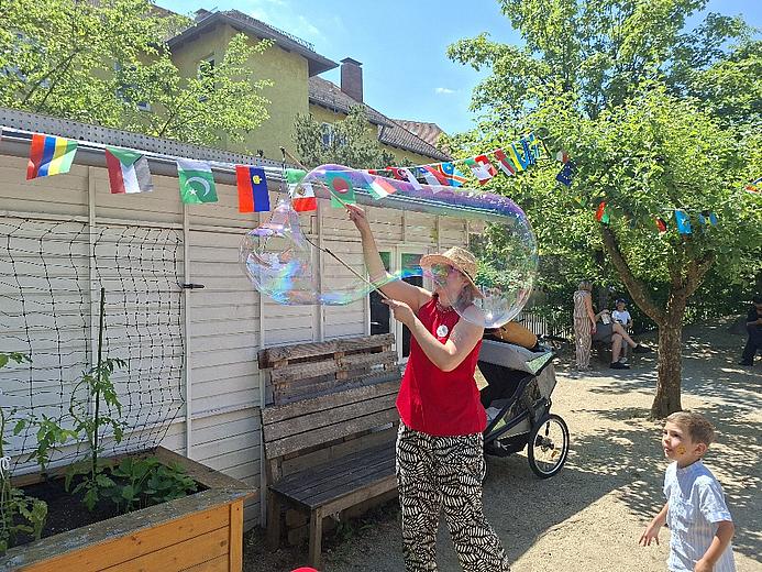 Eine Person erzeugt auf dem Sommerfest des Kindergartens mit Seifenblasenstäben eine große schillernde Seifenblase. Ein Kind beobachtet begeistert das Geschehen. Im Hintergrund sind bunte Länderflaggen, Bäume und weitere Besucherinnen und Besucher zu sehen.
