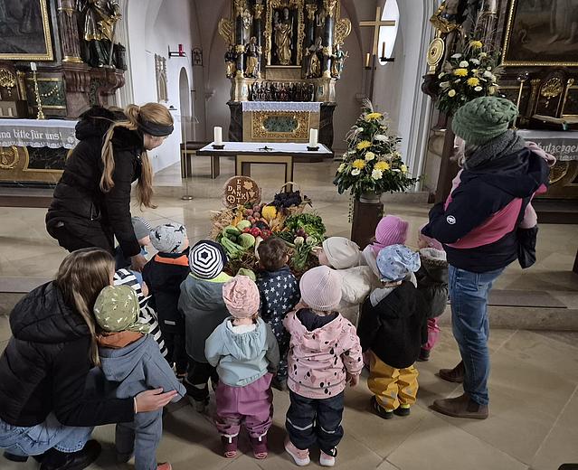 Mehrere kleine Kinder stehen gemeinsam mit zwei erwachsenen Begleitpersonen in einer Kirche vor einem Erntedankaltar. Auf dem Altarboden ist ein großer Korb mit buntem Obst, Gemüse, Getreide und Blumen zu sehen, geschmückt mit einem Schild, auf dem „Erntedank“ steht. Im Hintergrund erkennt man den festlich geschmückten Altarraum mit goldenen Verzierungen, Blumenarrangements und Kerzen. Die Kinder tragen bunte Jacken und Mützen und betrachten neugierig die Erntegaben. Die Stimmung wirkt ruhig und andächtig.