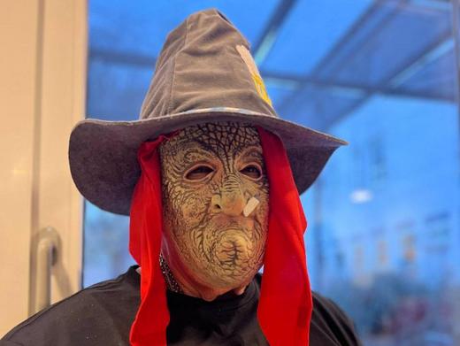 Das Bild wurde während der Faschingsfeier aufgenommen. Es zeigt eine Person, die eine Verkleidung trägt. Sie trägt eine Maske mit einem gealterten, runzeligen Gesicht, einen spitzen Hut und rote Stoffbänder statt Haare. Auf dem schwarzen T-Shirt steht die Aufschrift "Böser Alter" und ein Totenkopf. Im Hintergrund ist ein Fenster zu sehen.