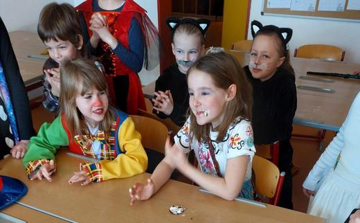 Das Bild zeigt eine Gruppe von Kindern des Kinderhortes Nittendorf bei der Faschingsfeier. Die Kinder sind verkleidet. Zwei Mädchen sitzen an einem Holztisch, während andere Kinder um die beiden herumstehen und lachen. Eines der Mädchen trägt ein buntes Kostüm, ähnlich einer Clownverkleidung. Das andere Mädchen trägt ein helles, gemustertes Oberteil. Im Hintergrund sind weitere Kinder zu sehen, einige mit Katzenohren und aufgemalten Schnurhaaren. Die Stimmung wirkt fröhlich. 